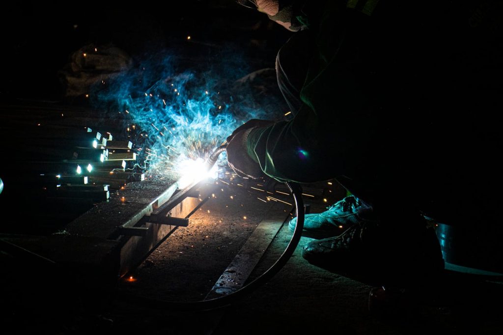 A welder works intensely with sparks flying, illuminating the dark workshop.